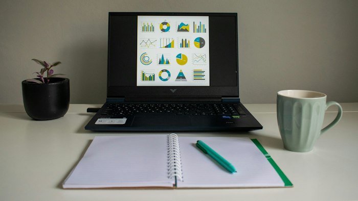 A laptop showing charts beside an open notebook and a mug on a bright desk