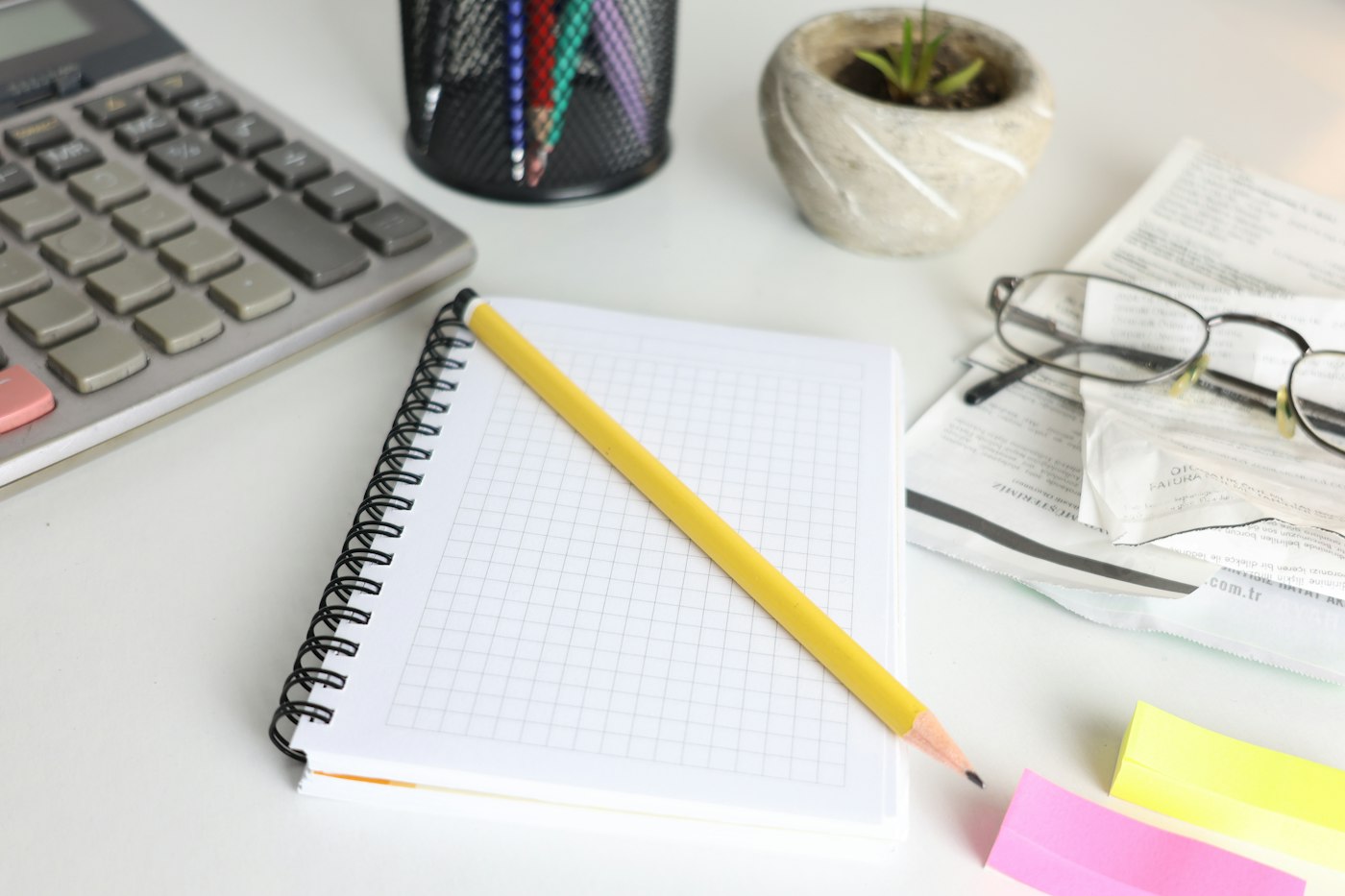 A notebook, a pencil, a calculator and reading glasses on a white desk